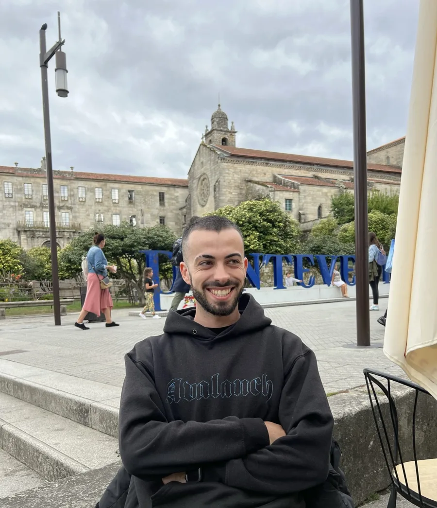 Portrait of Daniel Chazarra, smiling, wearing a black hoodie, seated outdoors in a Spanish town square